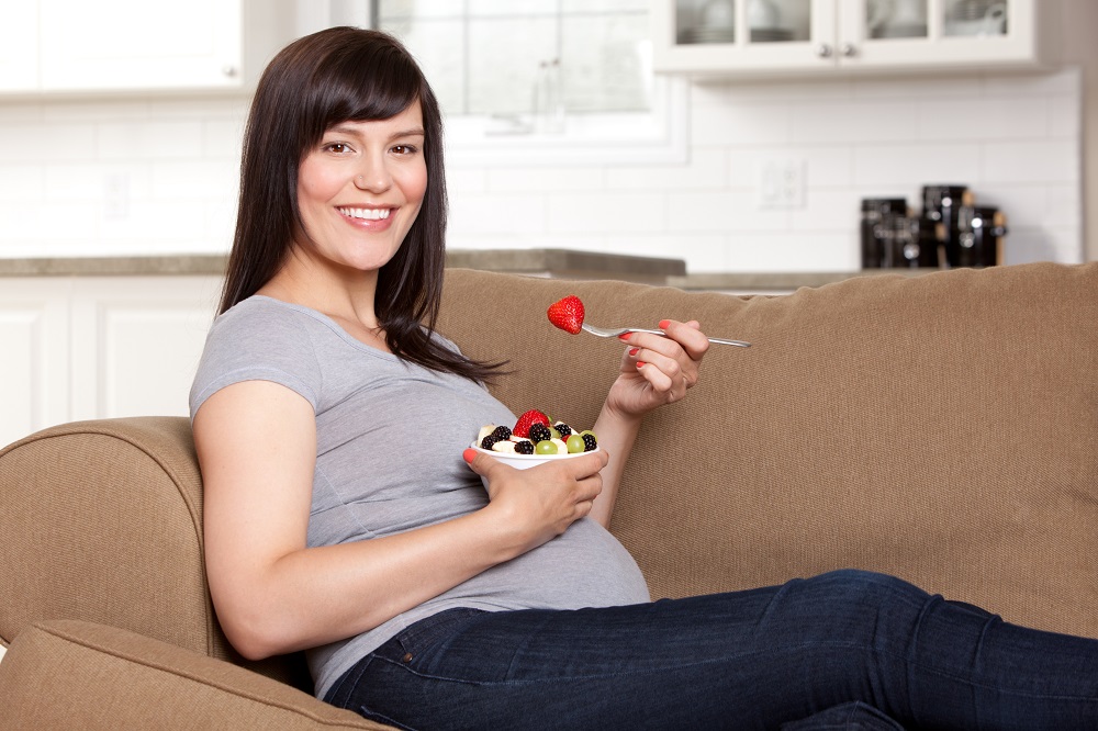Happy pregnant woman sitting on a sofa eating bowl of fresh fruit